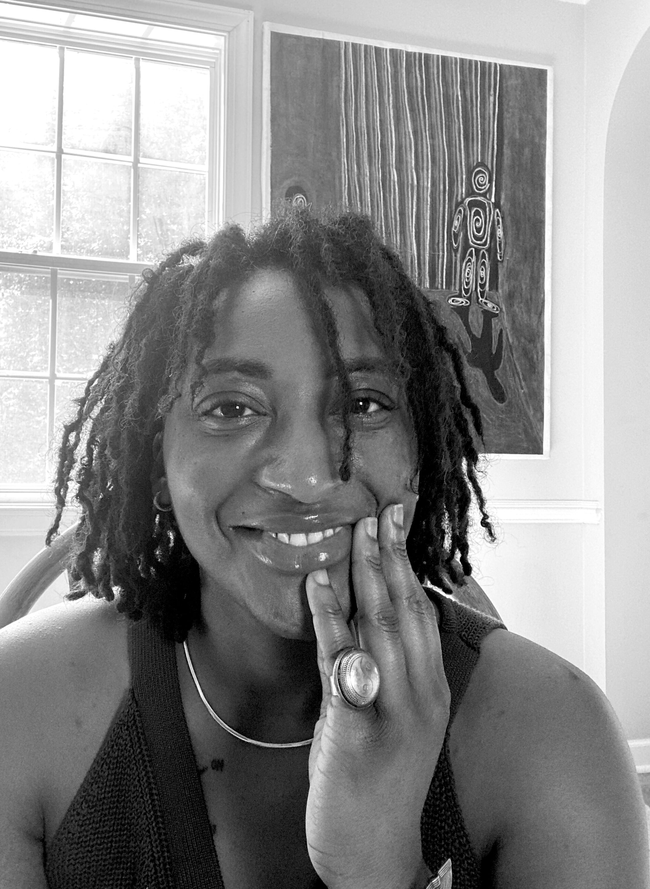 Person with dreadlocks and a ring, sitting indoors with a window and artwork in the background.